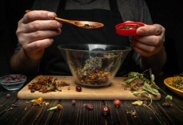 Herbalist skillfully prepares medicinal tea with berries and dry herbs. Hand adds dry carnation with a spoon to a glass bowl. Concept of traditional medicine at home.